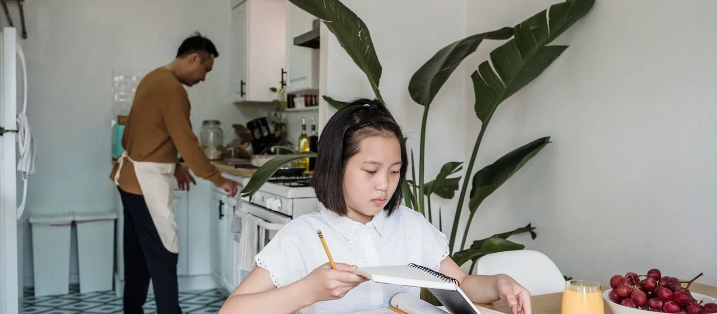 A child studying independently at home while a parent works nearby